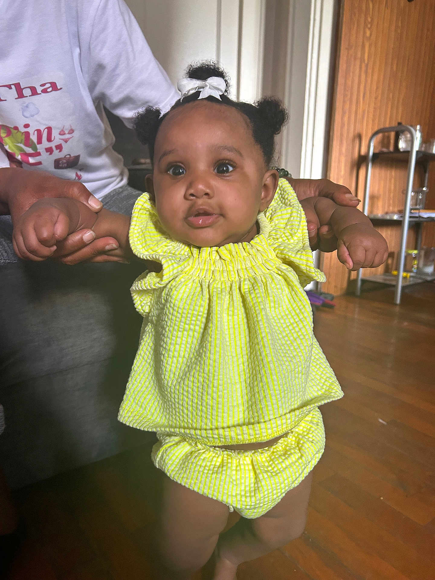 Aziaha joined the competition — help win amazing prizes! baby, child, infant, yellow_outfit, hair_bow, indoor, wooden_floor, holding_hands, curious_expression, person, adult_hands, bright_clothing, standing, home_interior, flooring, furniture, portrait, cute, small_child, people