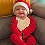 baby, child, santa_claus_outfit, red_clothing, hat, smile, face, couch, blanket, indoors, holiday, festive, cute, warm_clothing, sitting, young_child, cozy, home, person, portrait