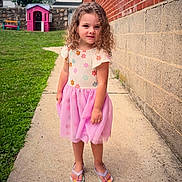 Myla is registered to the contest to win money with this photo: child, girl, curly_hair, floral_dress, pink_skirt, flip_flops, sidewalk, brick_wall, stone_wall, grass, playhouse, bench, outdoor, smiling, person, daylight, summer, cute, portrait, young