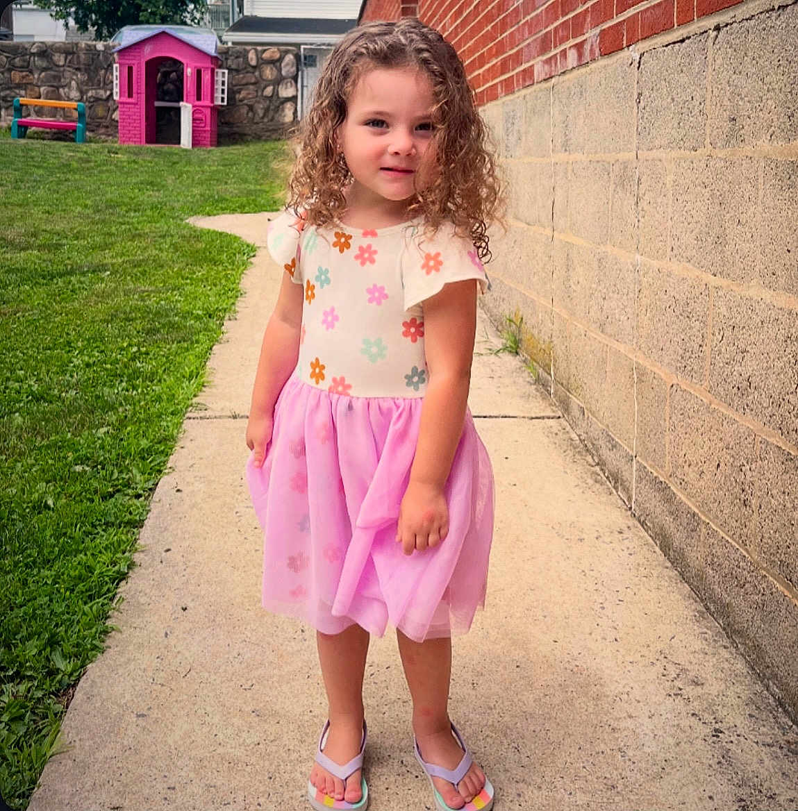 Myla is registered to the contest to win money with this photo: child, girl, curly_hair, floral_dress, pink_skirt, flip_flops, sidewalk, brick_wall, stone_wall, grass, playhouse, bench, outdoor, smiling, person, daylight, summer, cute, portrait, young