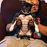 beige_pants, black_tshirt, brown_skin, casual_clothing, costume, couch, cowboy_hat, decor, dog, funny, glasses, holding, indoor, man, muscular_costume, person, pet, relaxing, short_hair, smiling