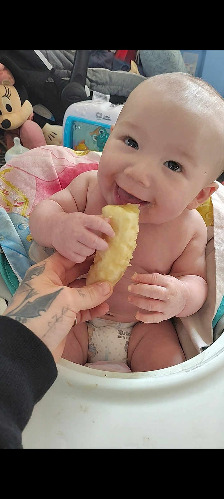 Greyson is registered to the contest to win money with this photo: baby, child, high_chair, food, hand, tattoo, smile, happy, toy, stuffed_animal, minnie_mouse, blanket, diaper, indoor, face, person, feeding, cute, infant, playful