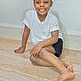 barefoot, black_shorts, casual_clothing, child, cute, face, feet, floor, hand, happy, indoors, person, portrait, sitting, skin, smiling, wall, white_tshirt, wooden_floor, young