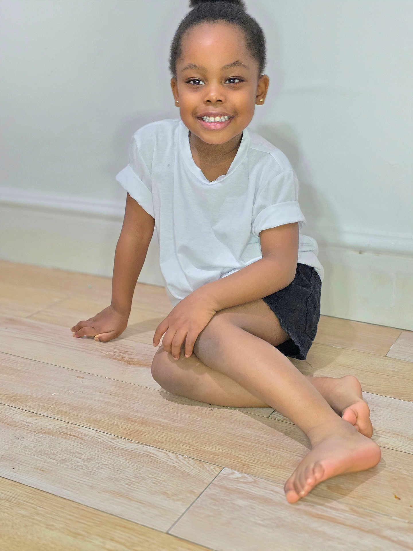 Chioma is registered to the contest to win money with this photo: barefoot, black_shorts, casual_clothing, child, cute, face, feet, floor, hand, happy, indoors, person, portrait, sitting, skin, smiling, wall, white_tshirt, wooden_floor, young