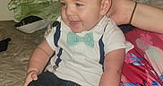 Owen is registered to the contest to win money with this photo: adult_arm, baby, bed, bedroom, blanket, bow_tie, child, clothing, cozy, happy, indoor, infant, jeans, pillow, portrait, shirt, sitting, smiling, sneakers, suspenders