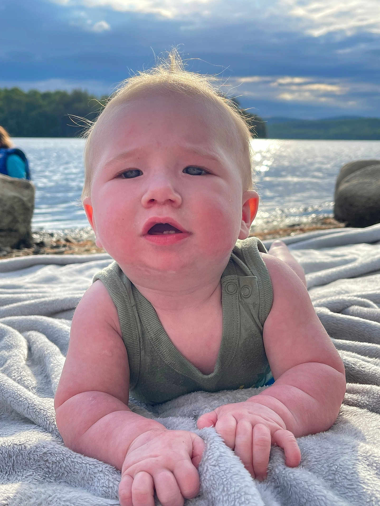 Grayson joined the competition — help win amazing prizes! baby, infant, blanket, water, lake, sky, clouds, sunlight, nature, outdoor, child, face, arms, expression, curious, green_clothing, rocks, scenery, portrait, relaxing