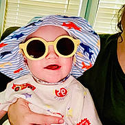 Anakyn is registered to the contest to win money with this photo: baby, child, sunglasses, hat, hand, person, indoor, window_blinds, smile, tshirt, colorful, patterned_clothing, adult_hand, seated, cheerful, young_child, casual, portrait, infant, holding