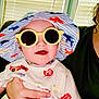 baby, child, sunglasses, hat, hand, person, indoor, window_blinds, smile, tshirt, colorful, patterned_clothing, adult_hand, seated, cheerful, young_child, casual, portrait, infant, holding