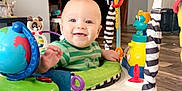 Anakyn joined the competition — help win amazing prizes! baby, child, infant, smiling, activity_jumper, toys, indoor, wooden_floor, green_shirt, playful, colorful, happy, face, foot, seat, home, furniture, person, cute, bright