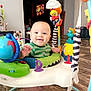 baby, child, infant, smiling, activity_jumper, toys, indoor, wooden_floor, green_shirt, playful, colorful, happy, face, foot, seat, home, furniture, person, cute, bright