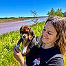 Ronnie is registered to the contest to win money with this photo: woman, puppy, dog, beagle, smile, outdoor, river, grass, blue_sky, sunny, nature, person, holding, happy, casual_clothing, tattoo, greenery, portrait, daylight, animal