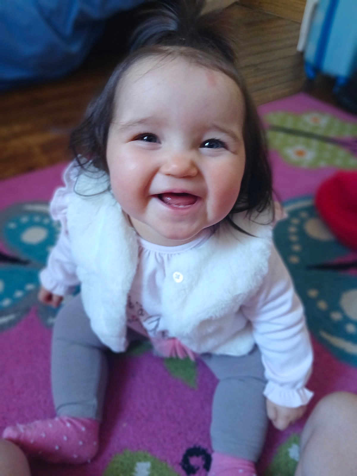 Inaya a rejoint le concours — aidez-le/la à gagner de superbes lots ! baby, child, smile, happy, sitting, pink_clothing, gray_pants, white_vest, carpet, indoor, person, cute, portrait, young_child, female, top_ponytail, sock, floor, home, playful