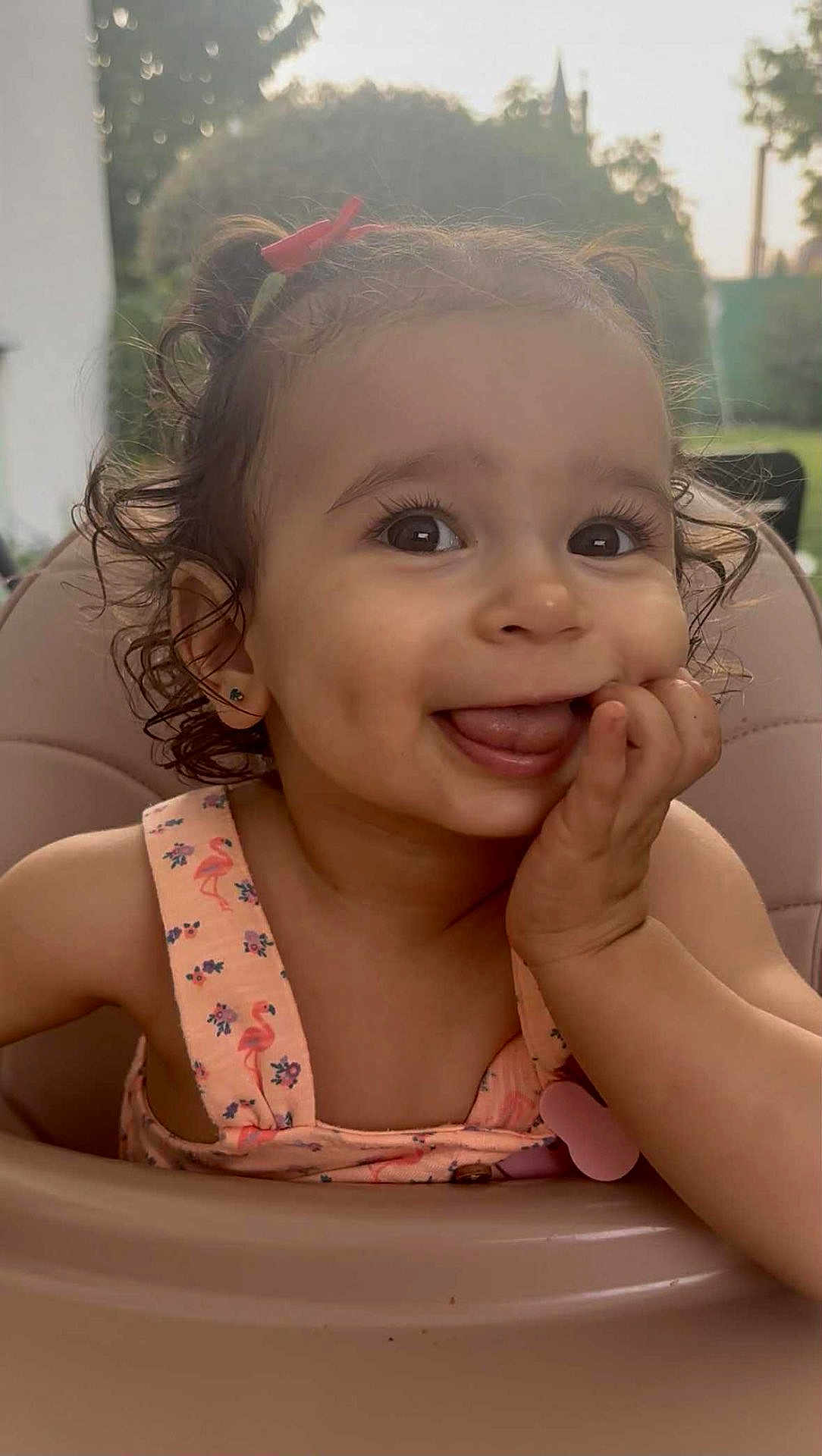 Asma participe au concours pour gagner de l'argent avec cette photo : baby, child, close_up, curly_hair, cute, daylight, earrings, flamingo_pattern, funny_face, hand_on_cheek, happy, high_chair, outdoor, person, pink_dress, portrait, seat, smiling, toddler, tongue_out