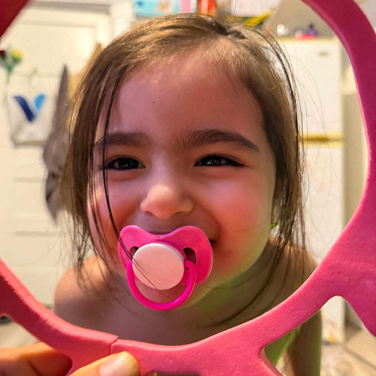 Hanna is registered to the contest to win money with this photo: child, toddler, portrait, smiling, pacifier, pink, toy, toy_frame, face, eyes, hair, indoor, home, playtime, close_up, hand, foreground, colorful, happy, family
