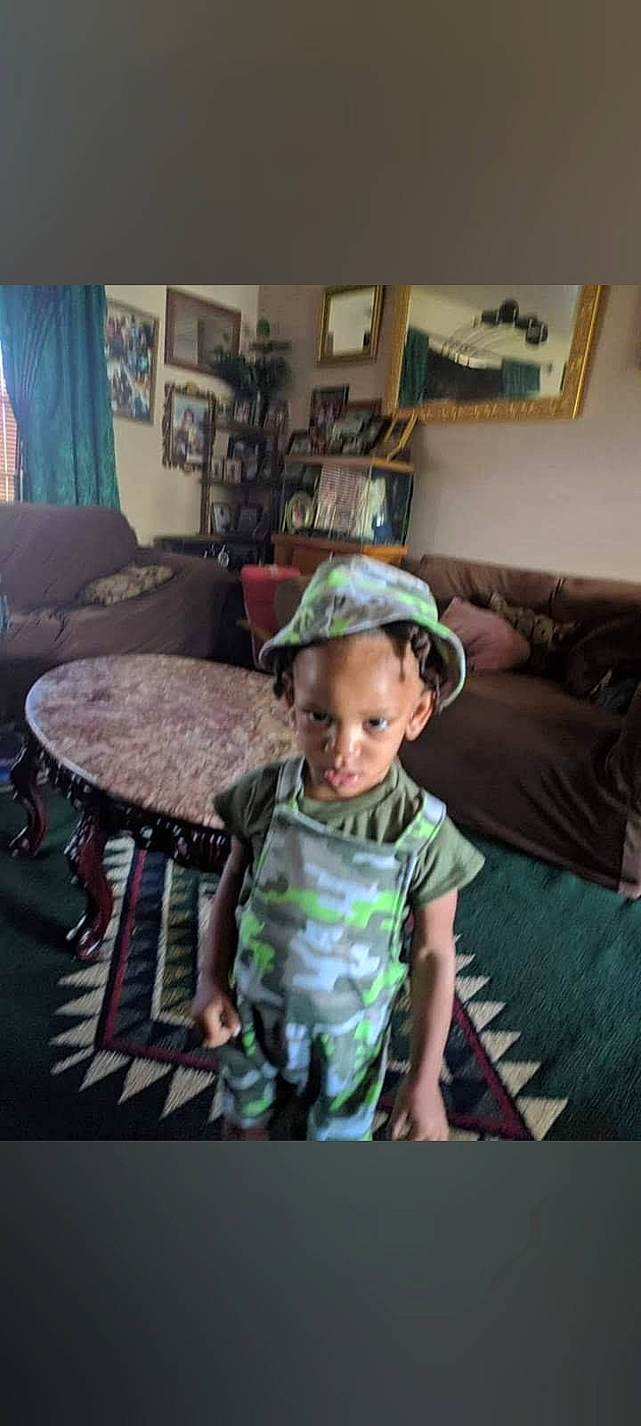 Devodric Ty'Shun is registered to the contest to win money with this photo: baseball_cap, bookcase, cap, comfort, couch, curtain, electronic_device, flooring, happy, hat, living_room, person, picture_frame, room, shelf, sitting, sleeve, t_shirt, table, toddler