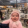baby, infant, smiling_baby, pink_hood, tiara, fluffy_jacket, shopping_store, retail_shelves, sock_display, hand_holding, happy_expression, chubby_cheeks, zipper, store_lighting, accessory_display, handbags, products, aisle, portrait, cute