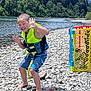 child, life_jacket, river, rocks, trees, outdoor, playpen, water, nature, summer, barefoot, blue_shorts, play, smile, forest, recreation, sunny, fun, adventure, person