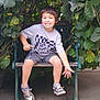 child, boy, smile, bench, leafy_plant, greenery, outdoor, happy, casual_clothing, sneakers, shorts, tshirt, nature, foliage, young, person, daylight, concrete, seated, pointing