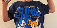 Ollie joined the competition — help win amazing prizes! child, boy, pokemon_card, sonic_tshirt, shorts, smiling, face_paint, peace_sign, indoor, wall, cushion, seated, black_hair, casual_clothing, playful, young, portrait, happy, hand_gesture, person