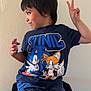 Ollie is registered to the contest to win money with this photo: boy, casual_clothing, child, cute, hand, indoor, peace_sign, person, playful, portrait, pose, short_hair, shorts, sitting, skin, smile, sonic, tshirt, wall, young