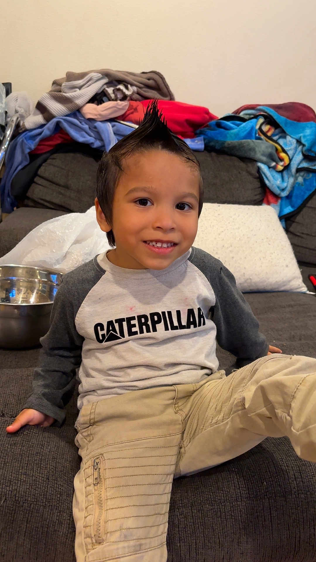 Ollie is registered to the contest to win money with this photo: child, boy, smile, mohawk, caterpillar_shirt, pants, couch, blankets, metal_bowl, indoor, casual_clothing, happy, young, portrait, seated, living_room, relaxed, cute, playful, home