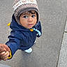 child, toddler, winter_hat, jacket, hand_holding, sidewalk, pavement, outdoor, curious, person, clothing, footwear, urban, cold_weather, looking_up, portrait, casual, walking, baby, cute