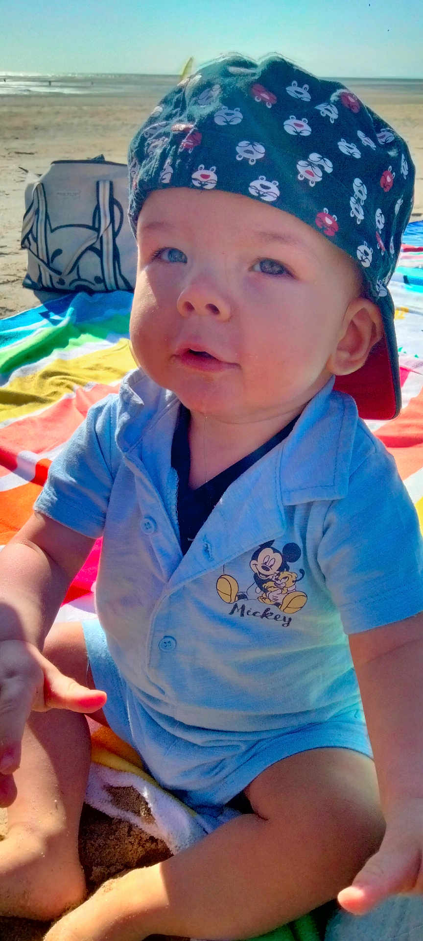 Marvynn a rejoint le concours — aidez-le/la à gagner de superbes lots ! baby, child, beach, towel, cap, hat, sand, sunlight, blue_shirt, mickey_mouse, cartoon, outdoor, summer, portrait, cute, sitting, toy, playful, vacation, happy