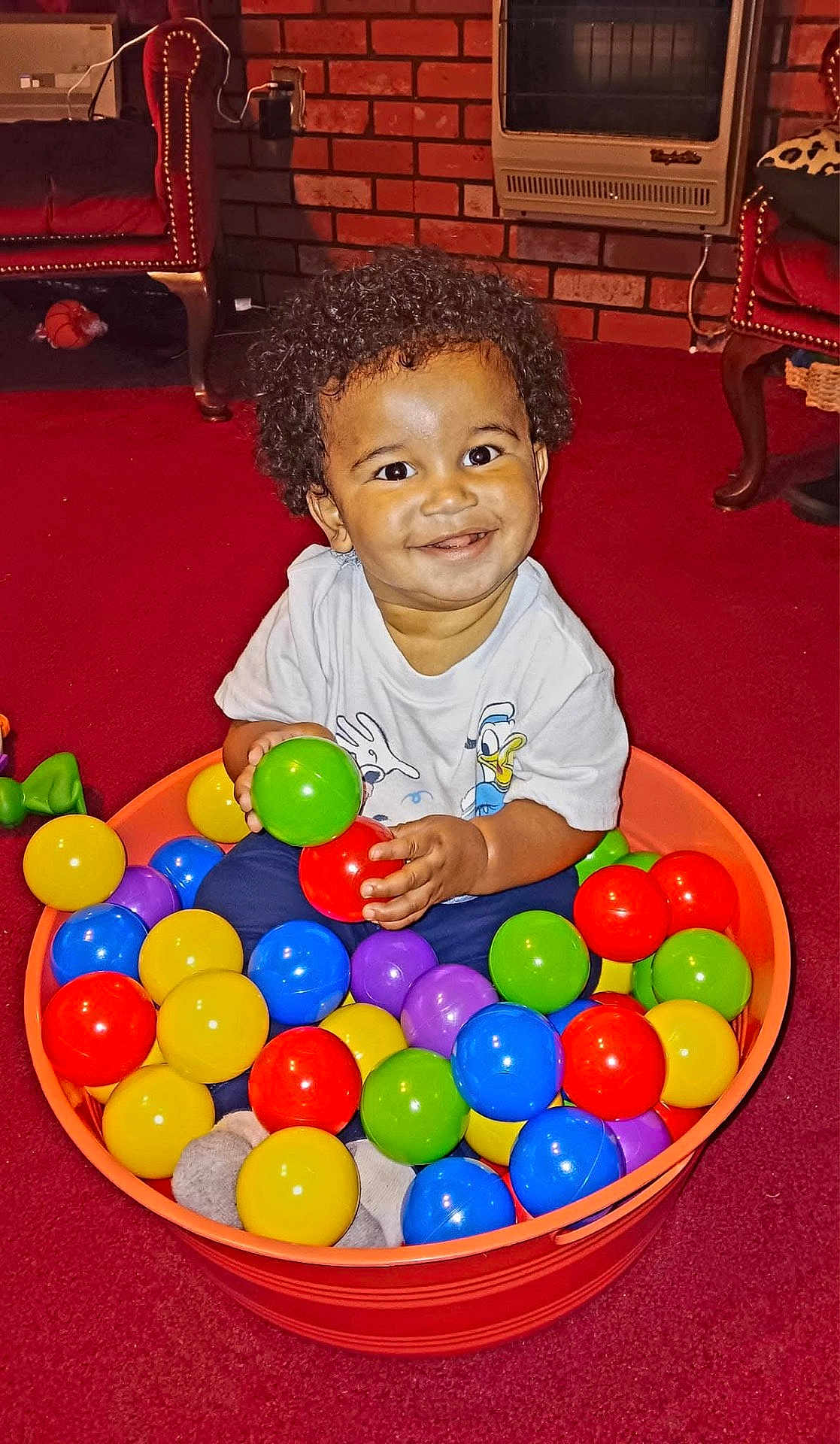 Silas is registered to the contest to win money with this photo: toddler, child, smiling, curly_hair, bucket, colorful_balls, red_carpet, indoor, toy, play, happy, person, sitting, plastic_balls, orange_bucket, blue_pants, white_shirt, furniture, heater, home