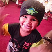 Silas is registered to the contest to win money with this photo: toddler, child, smiling, hat, beanie, dinosaur_patch, black_clothing, motion_blur, indoor, carpet, living_room, red_carpet, furniture, chair, holiday_decor, christmas_tree, happy, cute, portrait, person