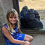 child, girl, blue_clothing, sportswear, smile, crouching, glass_window, gorillas, wildlife, animal_group, reflection, outdoor, nature, sand, tree, bracelet, footwear, human_animal_interaction, zoo, enclosure
