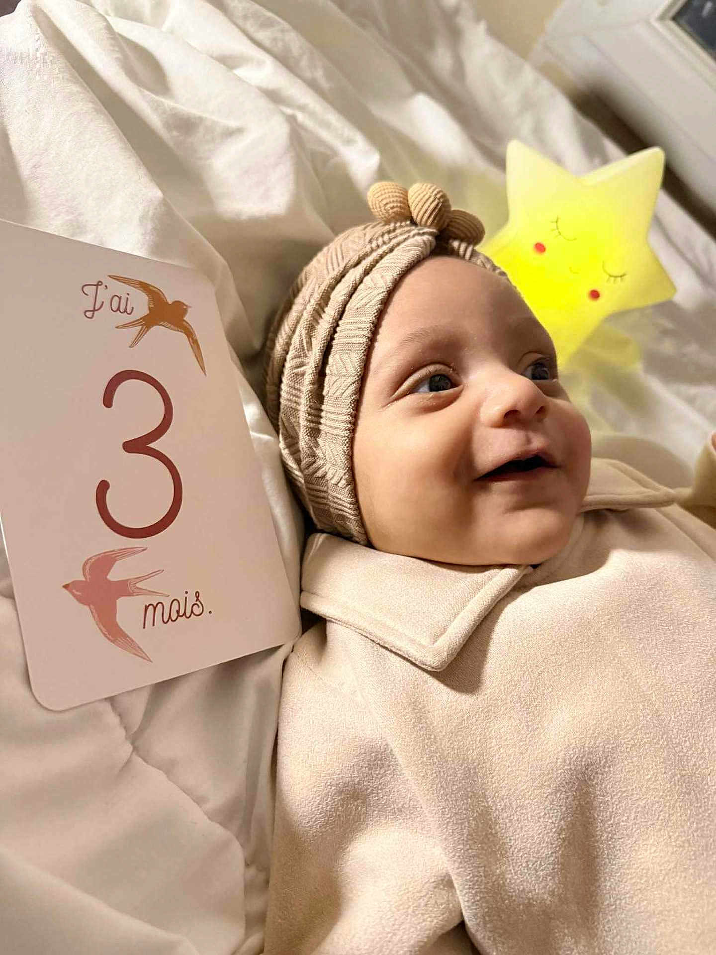 Zel a rejoint le concours — aidez-le/la à gagner de superbes lots ! baby, infant, smile, milestone_card, 3_months, beige_clothing, turban, blanket, bedroom, star_nightlight, soft_texture, cute, lying_down, portrait, indoors, happy, face, child, newborn, cozy