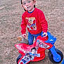 blue_jeans, boy, cap, casual_clothing, child, daytime, grass, happy, outdoor, pacifier, park, paw_patrol, person, playground, red_sweatshirt, smile, toy, toy_bike, vehicle, young_child
