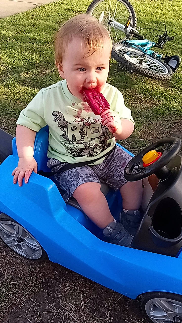 Jedidiah is registered to the contest to win money with this photo: baby_toddler_clothing, blue, bumper, child, cool, finger, fun, grass, hairstyle, happy, leisure, motor_vehicle, person, product, riding_toy, shorts, smile, tire, toddler, vehicle