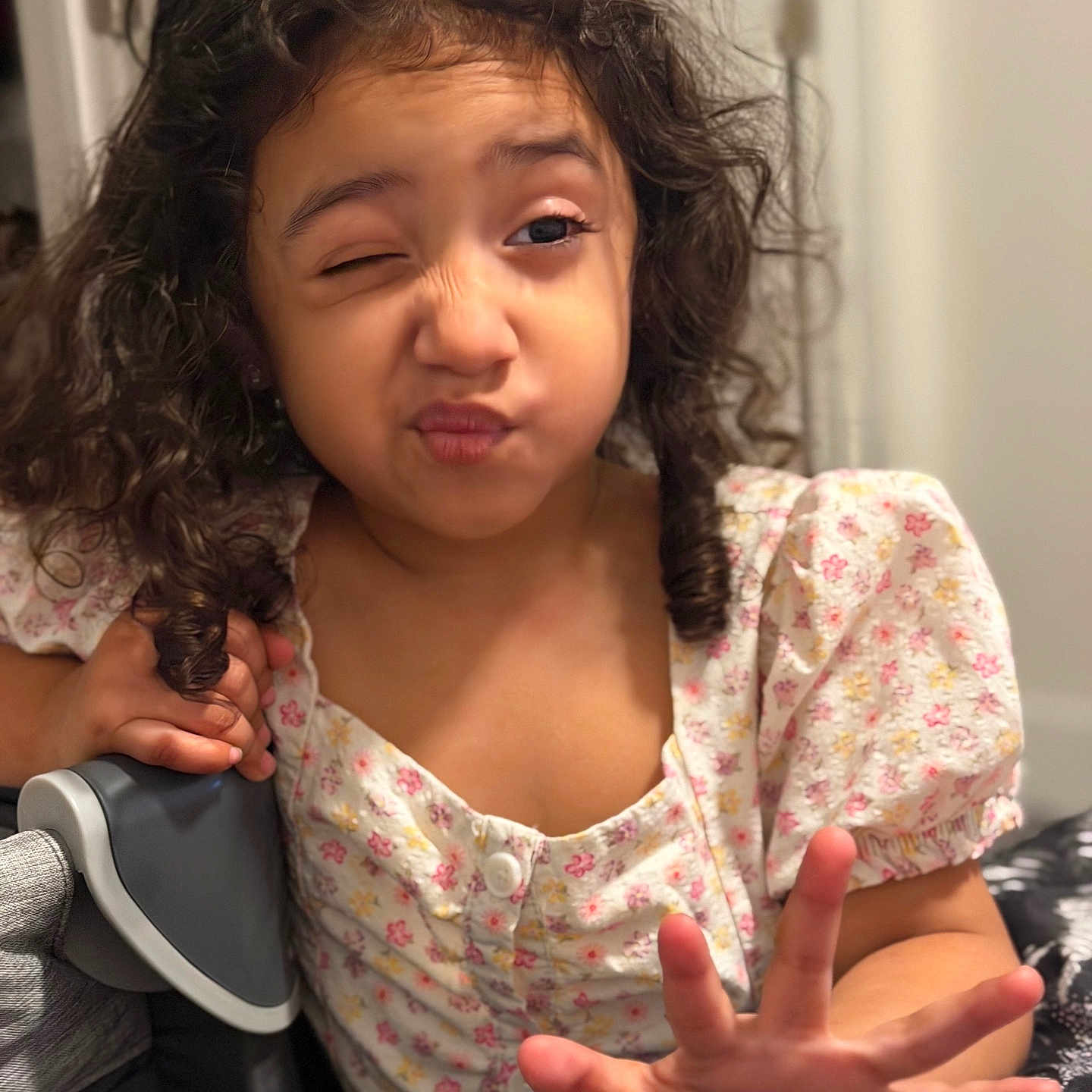 Nicole is registered to the contest to win money with this photo: child, girl, portrait, winking, funny_face, curly_hair, long_hair, floral_dress, smiling, hand_gesture, open_hand, indoor, seated, chair, close_up, face, eyes, playful, home, photography