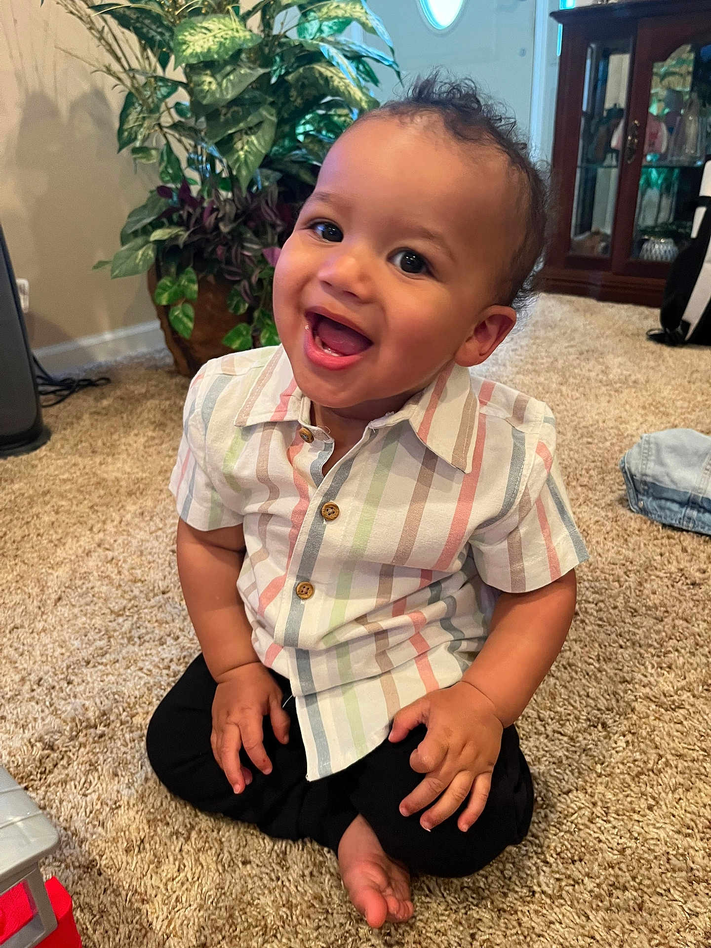 Zayden is registered to the contest to win money with this photo: toddler, child, smiling, happy, indoor, carpet, striped_shirt, black_pants, plant, home, furniture, cabinet, door, barefoot, cute, portrait, person, sitting, young_child, curly_hair