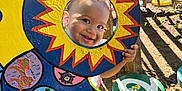 Zayden is registered to the contest to win money with this photo: child, toddler, face, smile, outdoor, sunlight, playground, wood, colorful, painting, cutout, hand_painted, garden, shadow, nature, happy, play, art, sign, fence