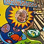 child, toddler, face, smile, outdoor, sunlight, playground, wood, colorful, painting, cutout, hand_painted, garden, shadow, nature, happy, play, art, sign, fence