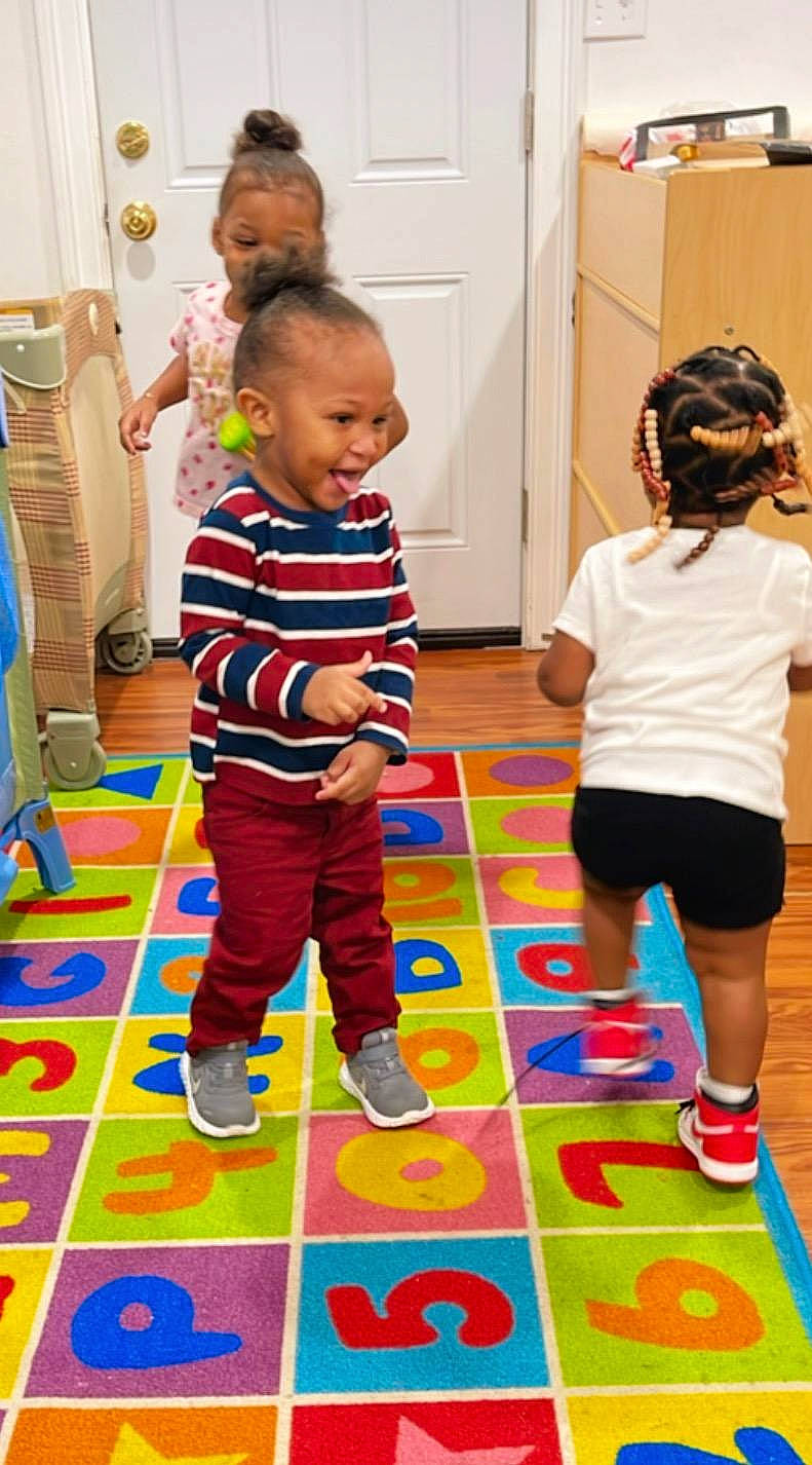 Micah is registered to the contest to win money with this photo: baby, child, door, event, floor, flooring, fun, joint, leg, leisure, mammal, pattern, person, purple, recreation, shorts, smile, standing, toddler, vertebrate
