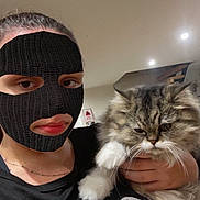 Pestouille a rejoint le concours — aidez-le/la à gagner de superbes lots ! cat, long_hair_cat, fluffy, fur, pet, animal, person, masked_person, selfie, portrait, indoors, ceiling_lights, black_shirt, text_on_clothing, whiskers, white_paws, gray_fur, holding_pet, close_up, funny_expression