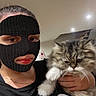 cat, long_hair_cat, fluffy, fur, pet, animal, person, masked_person, selfie, portrait, indoors, ceiling_lights, black_shirt, text_on_clothing, whiskers, white_paws, gray_fur, holding_pet, close_up, funny_expression