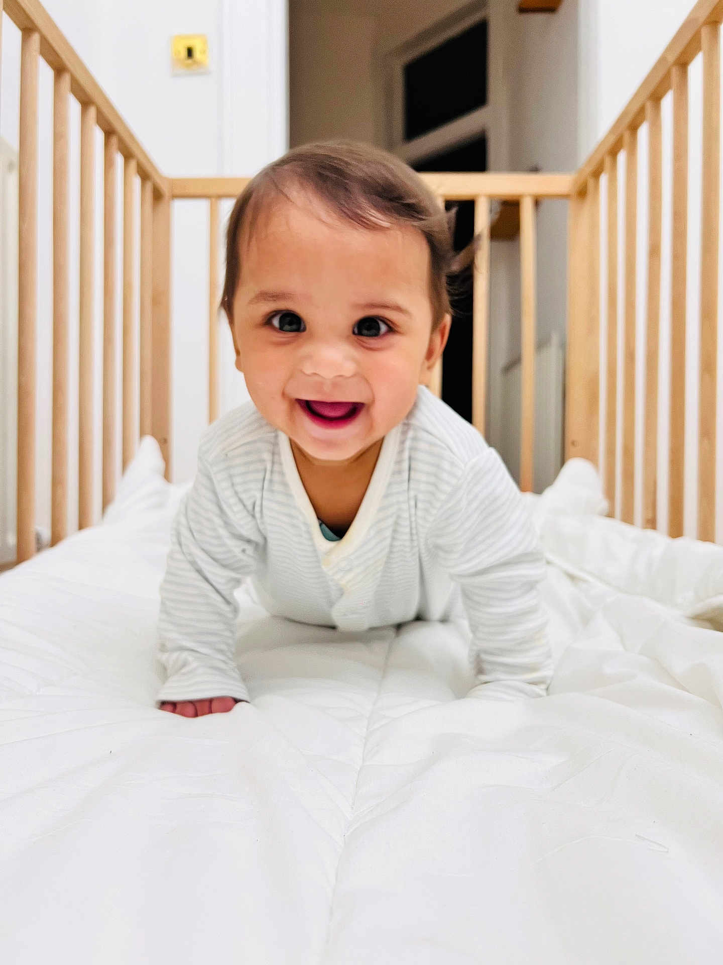 Arav joined the competition — help win amazing prizes! baby, infant, crib, crawling, smiling, happy, pajamas, wooden_crib, white_comforter, indoors, child, cute, face, portrait, bedroom, young_child, person, baby_clothes, playful, bright