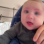 George is registered to the contest to win money with this photo: baby, child, face, hand, bracelet, bed, pillow, clothing, person, indoor, curious, portrait, gray_outfit, metal_bedframe, blue_pillow, open_mouth, close_up, human, skin, infant
