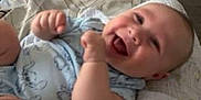 George is registered to the contest to win money with this photo: baby, infant, smiling, laughing, bed, blanket, patterned_sheet, clothing, indoor, furniture, happy, child, person, cute, cozy, soft_toys, room, relaxed, lying_down, skin