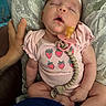 baby, sleeping, pacifier, pink_clothing, onesie, hand, lap, indoor, person, infant, cute, peaceful, relaxed, skin, furniture, pattern, holding, child, human, comfort