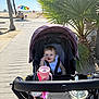 Zoe is registered to the contest to win money with this photo: baby, stroller, child, beach, boardwalk, sunny, umbrella, palm_tree, outdoor, drink, sippy_cup, happy, smiling, summer, daylight, person, seat, clothing, nature, vacation