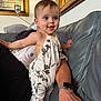 toddler, child, smiling, floral_clothing, couch, living_room, adult_arm, smartwatch, painting, artwork, home_interior, happy, portrait, person, furniture, indoor, baby, supporting_hand, casual, relaxed