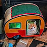 cat, cat_house, orange_cat, indoor, miniature_camper, pet, window, door, carpet, patterned_rug, cozy, fluffy, curious, toy, animal, home, furniture, decor, colorful, cute