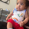 baby, child, person, indoor, clothing, red_shorts, sleeveless_top, sitting, hand, face, wide_eyes, expression, adult_hand, furniture, painting, shelf, room, sock, comfort, portrait