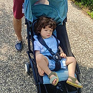 Anas a rejoint le concours — aidez-le/la à gagner de superbes lots ! toddler, child, stroller, curly_hair, blue_clothing, sandal, adult, hand, watch, gravel_path, greenery, flowers, outdoor, sunny, casual_clothing, footwear, person, nature, walking, daylight