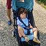 toddler, child, stroller, curly_hair, blue_clothing, sandal, adult, hand, watch, gravel_path, greenery, flowers, outdoor, sunny, casual_clothing, footwear, person, nature, walking, daylight